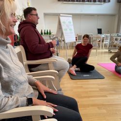 Menschen sitzen und dehnen sich auf Matten in einem hellen Raum mit einem Flipchart im Hintergrund.
