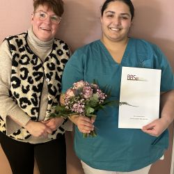 Zwei lächelnde Frauen halten ein Zertifikat und einen Blumenstrauß in einem hellen Raum.