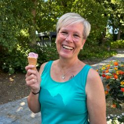 Eine lachende Frau im türkisfarbenen Top genießt ein Eis in einem sonnigen Park mit blühenden Blumen.