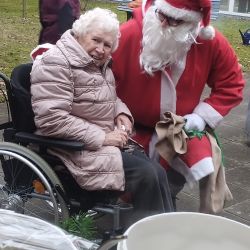 Ein älterer Mann im Weihnachtsmann-Kostüm besucht eine lächelnde Frau im Rollstuhl im Freien und verbreitet Freude.