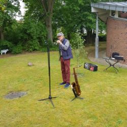 Ein Mann spielt Saxophon neben einem Mikrofonständer auf einer Wiese, umgeben von Bäumen und einem Backsteingebäude.