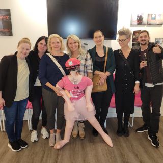 Eine Gruppe von Menschen posiert lachend mit einer aufblasbaren Puppe in einem Raum mit rosa Stühlen und Bildern an der Wand.