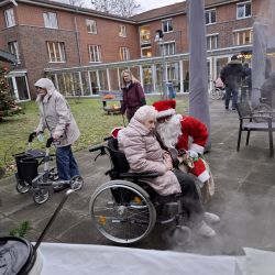 Ein älterer Mensch im Rollstuhl wird draußen vor einem festlichen Pflegeheim herzlich vom Weihnachtsmann umarmt.