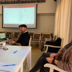 Ein Mann hält eine Präsentation vor einer Frau in einem Konferenzraum, während ein Bildschirm im Hintergrund leuchtet.