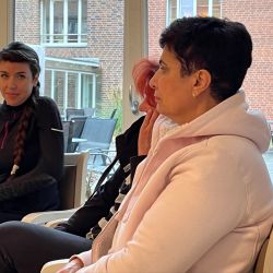 Drei Frauen sitzen in einem Raum mit großen Fenstern und unterhalten sich angeregt miteinander.