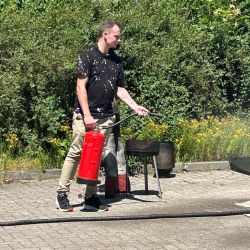 Ein Mann benutzt im Freien einen Feuerlöscher neben einem Grill auf einer gepflasterten Fläche.