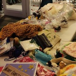 Auf einem Tisch liegen handgefertigte Weihnachtskarten und gestrickte Geschenke, während im Hintergrund zwei Personen sitzen.