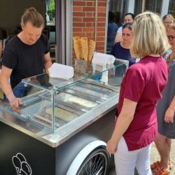 Eine Frau bestellt Eis an einem mobilen Eiswagen mit schwarz-weiß gestreiftem Dach vor einem Backsteingebäude.