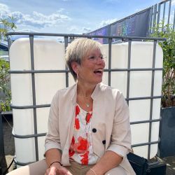 Eine lächelnde Frau in beiger Kleidung sitzt im Freien und genießt mit einem Glas den sonnigen Tag.