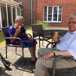 Ein älteres Paar sitzt auf einer Terrasse und genießt Eis, während ein Rollator neben ihnen steht.