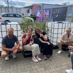 Vier Personen sitzen entspannt draußen auf "Honest"-Boxen und genießen gemeinsam ein Getränk unter bewölktem Himmel.