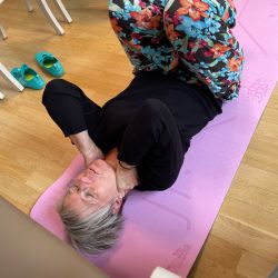 Eine Frau in bunten Blumenhosen macht auf einer lila Yogamatte eine Übung für den Nacken.