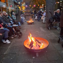 Menschen versammeln sich um Feuerstellen auf einem Innenhof, während Laternen den Abendhimmel schmücken.