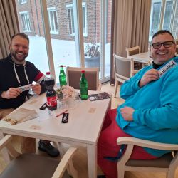 Zwei Männer sitzen lächelnd an einem Tisch und halten Schokoladenriegel in den Händen, während sie Getränke genießen.