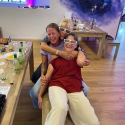 Drei lachende Frauen mit 3D-Brillen umarmen sich in einem Raum mit Weltraum-Wandbild, Getränke stehen auf dem Tisch.