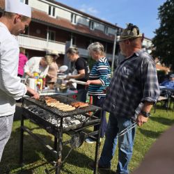 Ein Mann grillt Würstchen bei sonnigem Wetter, während fröhliche Menschen im Garten um den Grill versammelt sind.