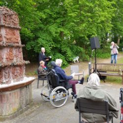 Ältere Menschen sitzen im Park nahe einem Brunnen und lauschen aufmerksam einem Vortrag im Freien.