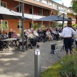 Ältere Menschen genießen ein Freiluftkonzert unter Sonnenschirmen vor einem charmanten Backsteingebäude.