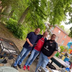 Drei Männer stehen lächelnd neben einem Grill im Freien, umgeben von Bäumen und einer Backsteinfassade.