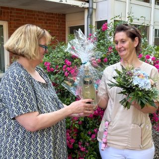 Zwei Frauen stehen lächelnd vor blühenden Sträuchern, während eine die andere mit einer Flasche und Blumen beschenkt.