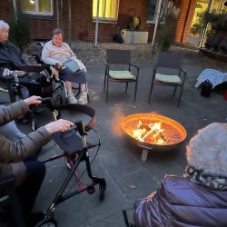 Senioren sitzen entspannt in Rollstühlen um eine Feuerstelle im Innenhof und genießen den gemütlichen Abend.