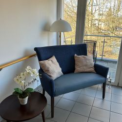 Ein gemütliches Sofa mit Kissen steht neben einem Tisch mit Orchidee und Lampe, die auf einen sonnigen Balkon blicken.