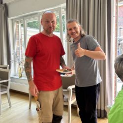 Zwei Männer stehen lachend in einem sonnigen Raum, einer hält fröhlich einen Teller mit Kuchen.