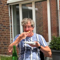 Ein Mann in gestreiftem T-Shirt genießt draußen ein Stück Pizza an einem Tisch mit einer Flasche Sprite.