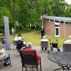 Ältere Menschen sitzen im Garten und hören einer Person in schwarzem Gewand bei einem Vortrag vor einem Gebäude zu.