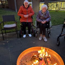 Zwei ältere Frauen im Rollstuhl genießen bei Abenddämmerung ein wärmendes Lagerfeuer im Garten vor einem Gebäude.