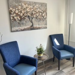 Zwei blaue Sessel flankieren einen runden Tisch mit Blumen, vor einem Gemälde eines blühenden Baumes an der Wand.