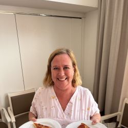 Eine lächelnde Frau in rosa Kleidung präsentiert stolz zwei Teller mit Pizza, Brot und Oliven in einem hellen Raum.