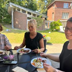 Drei Frauen genießen ein Mittagessen im Freien bei sonnigem Wetter vor einem charmanten Backsteingebäude.