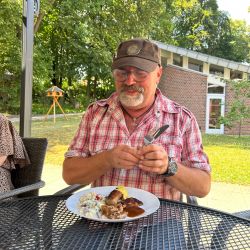Ein Mann in kariertem Hemd sitzt draußen und genießt ein Mittagessen mit Kartoffelsalat an einem Gartentisch.