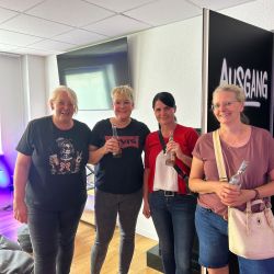 Vier Frauen stehen lächelnd mit Getränken in einem modernen Raum vor einem großen "Ausgang"-Schild zusammen.