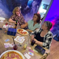 Drei Frauen genießen Pizzen an einem Tisch, während ein kosmisches Wandgemälde die Atmosphäre des Raumes bereichert.