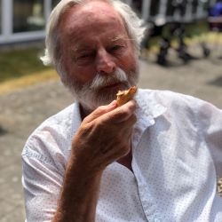 Ein älterer Mann mit weißem Bart genießt draußen entspannt ein Stück Pizza in der Sonne.