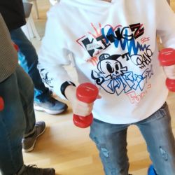 Ein Junge in sportlicher Kleidung hält lächelnd rote Gewichte in einem Raum voller spielender Kinder.