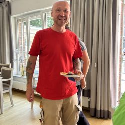 Ein Mann in rotem T-Shirt lächelt mit einem Teller Pizza in einem lichtdurchfluteten Raum.