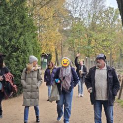 Eine Gruppe von Menschen spaziert durch einen herbstlichen Park, während eine Person eine lustige Tiermaske trägt.