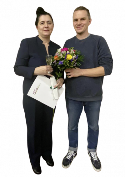 Eine Frau hält ein Zertifikat und ein Glas Sekt, während ein Mann ihr gratulierend Blumen überreicht.