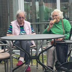 Zwei ältere Frauen sitzen draußen an einem Tisch, während eine von ihnen genüsslich ein Eis isst.