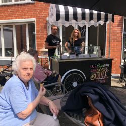 Eine ältere Frau genießt ein Eis auf einer sonnigen Terrasse vor einem mobilen Eiscreme-Stand mit fröhlichen Verkäufern.