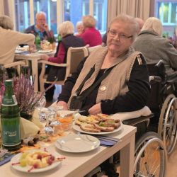 Eine ältere Frau im Rollstuhl genießt ein gemeinsames Essen in einem gemütlichen, lichtdurchfluteten Speisesaal.