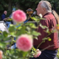 Ein Mann mit langen Haaren singt in ein Mikrofon, umgeben von Rosenblüten in einem malerischen Garten.