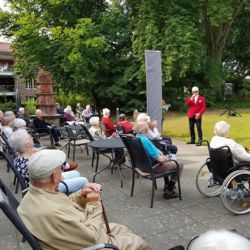 Ein Mann in rotem Sakko bietet Senioren im Garten eine musikalische Darbietung im Freien dar.