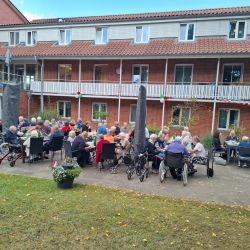 Ältere Menschen genießen ein geselliges Beisammensein im Freien, umgeben von bunten Girlanden vor einem roten Backsteingebäude.