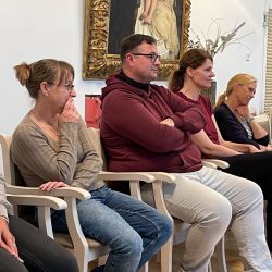 Eine Gruppe von fünf Personen sitzt aufmerksam nebeneinander in einem Raum und hört einem Vortrag zu.