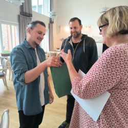 Drei Personen stehen in einem hellen Raum, während eine Frau einem Mann eine grüne Geschenktüte überreicht.