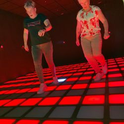 Zwei Personen tanzen fröhlich auf einem leuchtenden, rot und grün beleuchteten Boden und genießen den Moment.